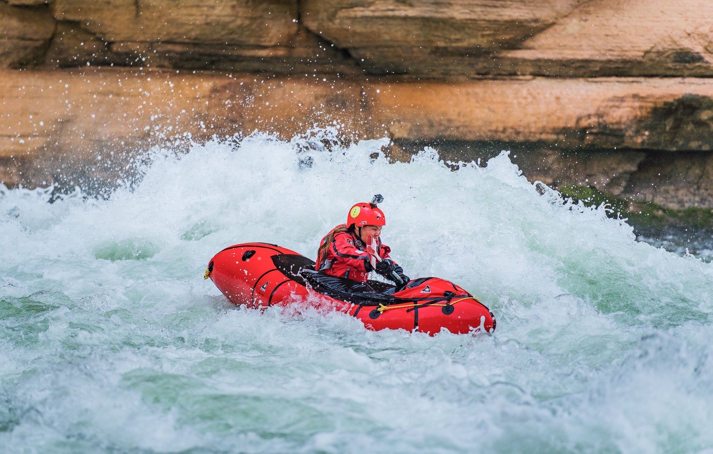Henkilö meloo punaista packraftia kuohuvassa koskessa kuivapuku päällä